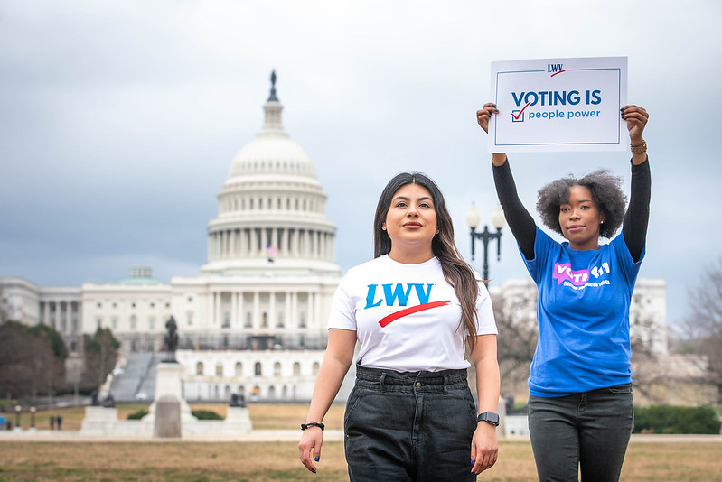 Two women standing in front of the US Capitol building. They are wearing LWV, VOTE411 t-shirts and holding signs.